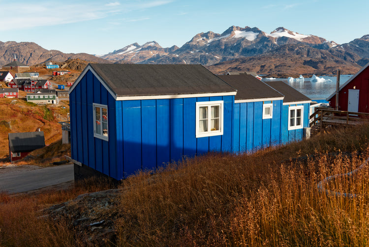 Blue wooden house on an autumn hillside, with icebergs and rugged mountains beyond in Tasiilaq, Greenland.
