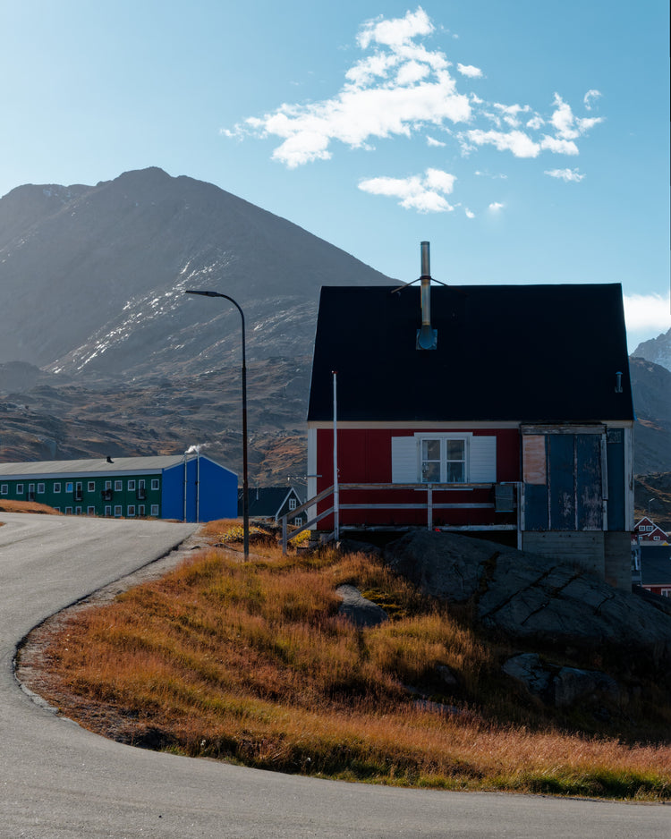 Red wooden house beside a curving road, with autumn grass and a mountain backdrop in Tasiilaq, Greenland.