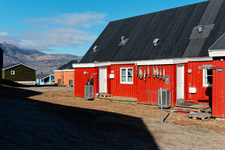 A red wooden house in Tasiilaq, Greenland, with fish hanging out to dry and deep shadow across the foreground.