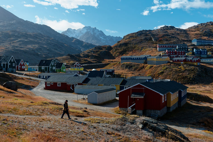 A walker crossing an autumn hillside above colourful houses, with rugged mountains beyond in Tasiilaq, Greenland.