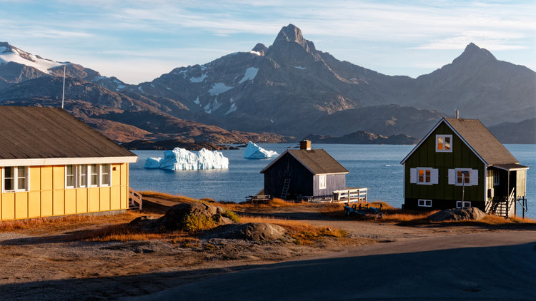 Yellow and green waterside houses with a small wooden house between them, white icebergs and rugged mountains in Tasiilaq, Greenland.