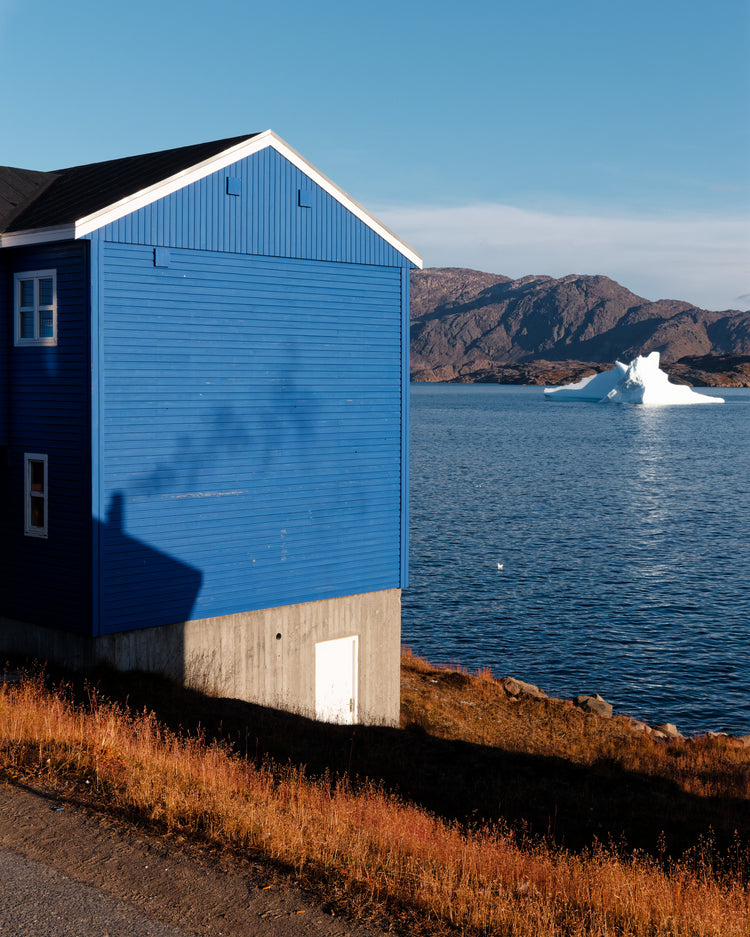 Blue wooden house beside the water, with a white iceberg, rocky mountains and autumn grass in Tasiilaq, Greenland.